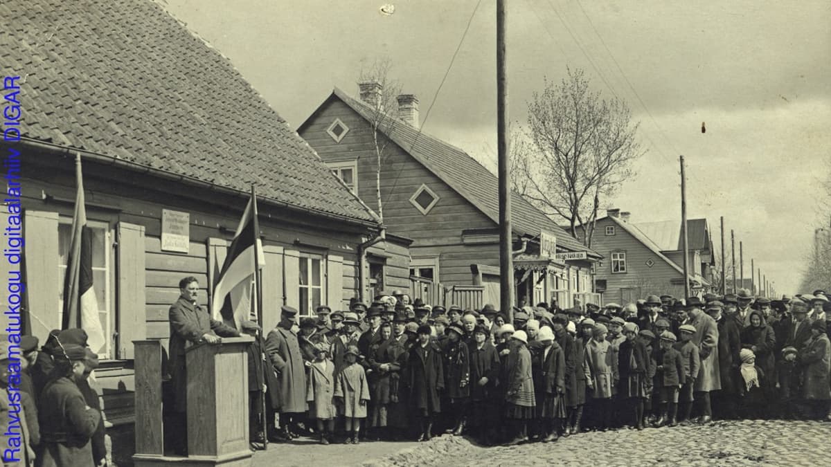 Mustavalkoinen valokuva väkijoukosta, joka seisoo kuuntelemassa puhujapöntössä olevaa miestä. Mukulakatu,puutaloja ja salossa Viron lippu.