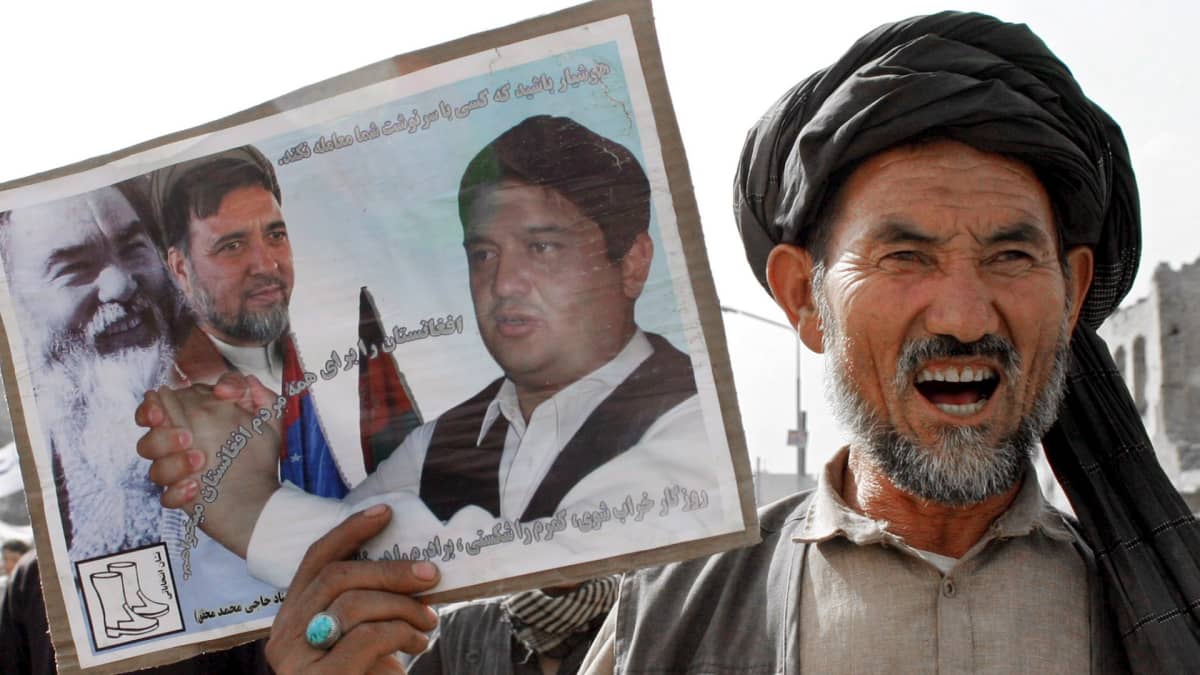 Kuvassa hazaramies pitää oikeassa kädessään taulua, jossa kolmen miehen valokuvat.