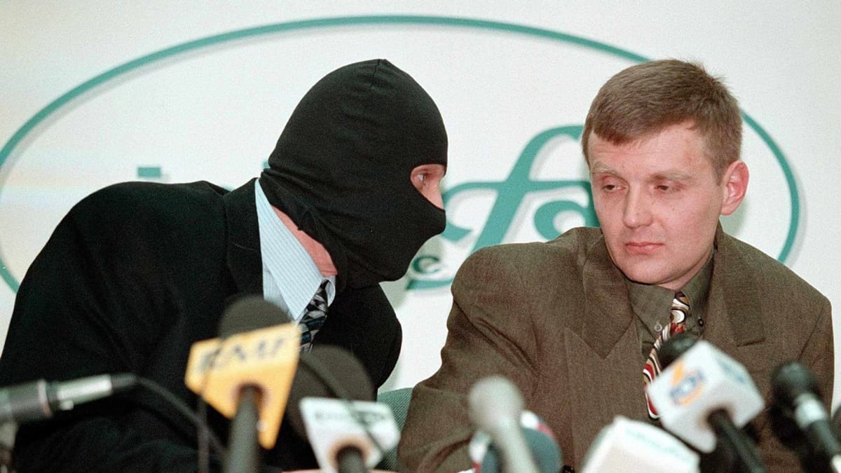 Mustaan hiihtomaskiin pukeutunut mies on kumartunut Litvinenkon puoleen. Litvinenkolla on ruskea puku ja vihreä kauluspaita sekä kirjava kravatti. Miehet istuvat pöydän takana. Pöydällä on mikrofoneja. Heidän takanaan seinällä näkyy uutistoimisto Interfaxin logo.