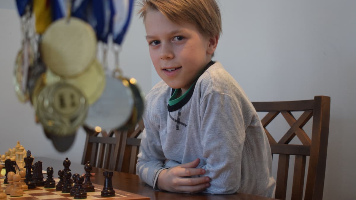 Poika istuu shakkipelilaudan ääressä ja kuvan edustalla roikkuu mitaleita