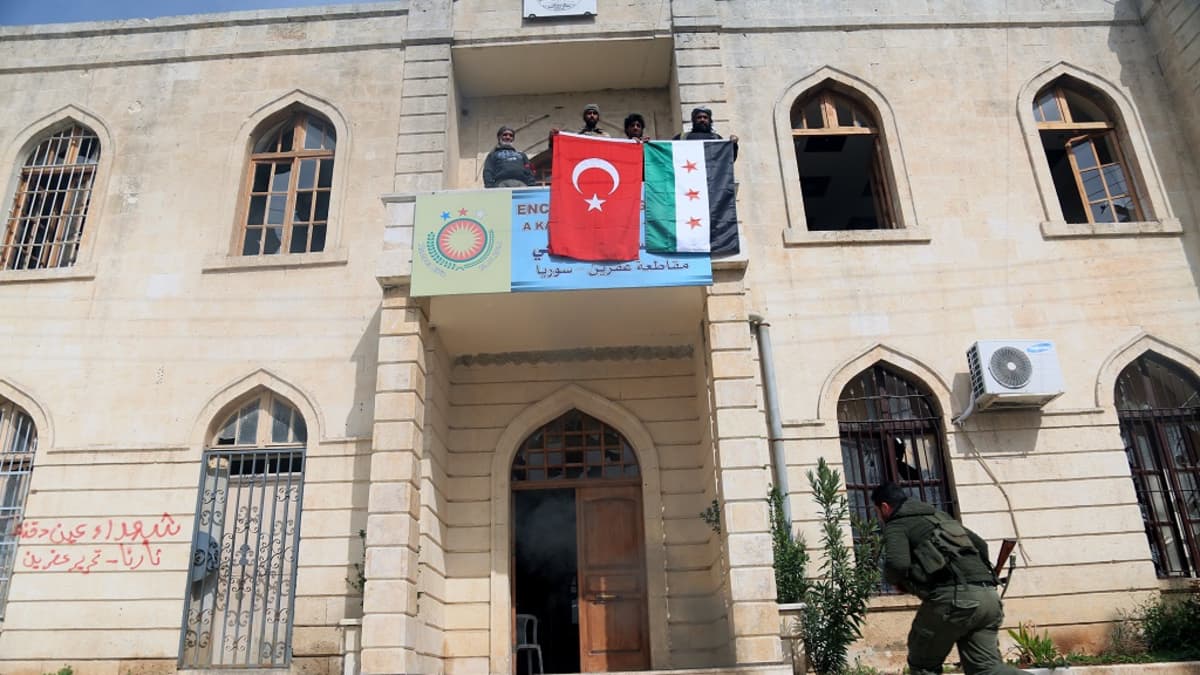 Mies juoksee kohti kivistä hallintorakennusta, jonka parvekkeelta miesjoukko roikuttaa Syyrian ja Turkin lippuja.