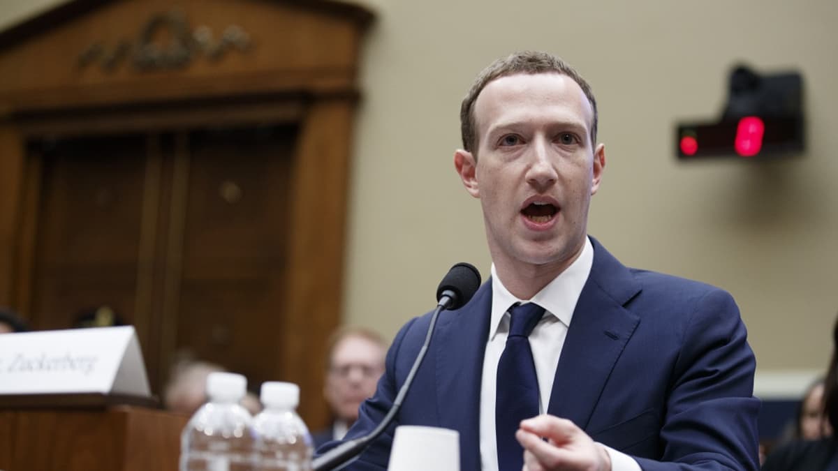 Tummansiniseen pukuun, valkoiseen kauluspaitaan ja tummansiniseen kravattiin pukeutunut Zuckerberg istuu pöydän takana ja puhuu mikrofoniin. Taustalla näkyy iso ovi. Pöydällä on kaksi vesipulloa.