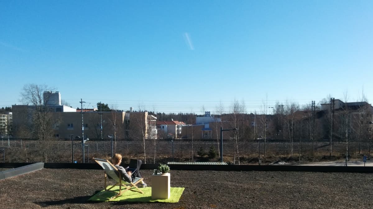 Opiskelija istuu rakennuksen tasakattoisella katolla aurinkotuolissa kannettava tietokone sylissä. Taustalla näkyy junarata ja kerrostaloja.