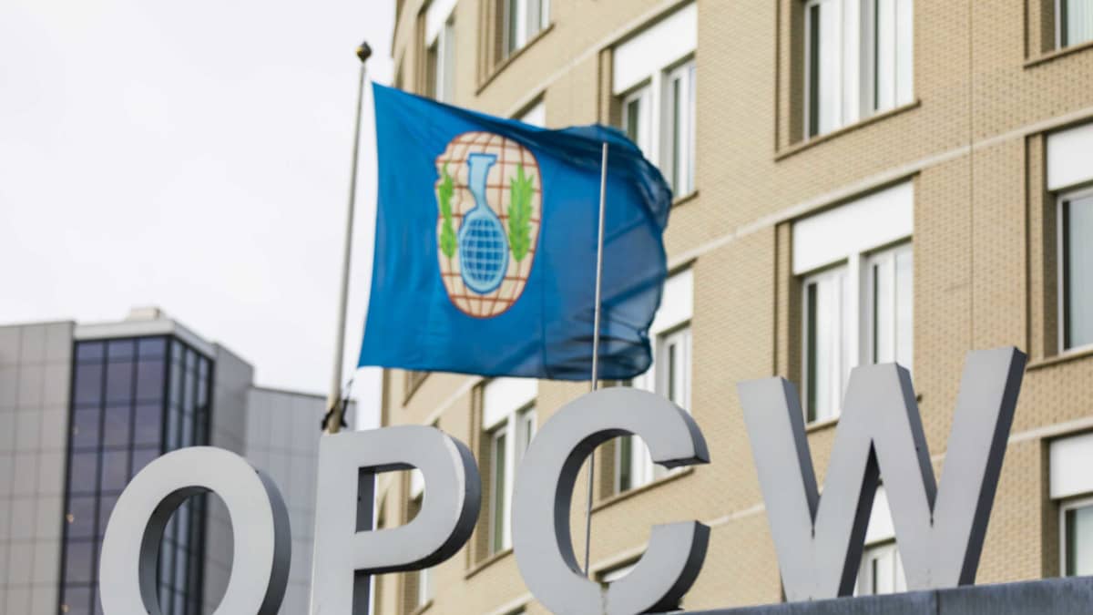 Kuvassa OPCW:n logo ja lippu, joka heiluu päämajan fasadissa.