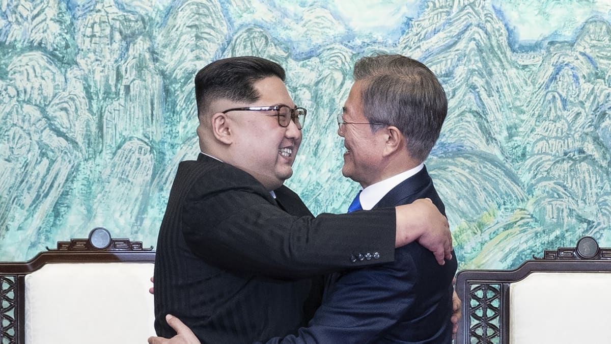 Kim ja Moon halaavat hymyillen kokoushuoneessa. Kimillä on musta maolaishenkinen puku ja Moonilla tummansininen puku.