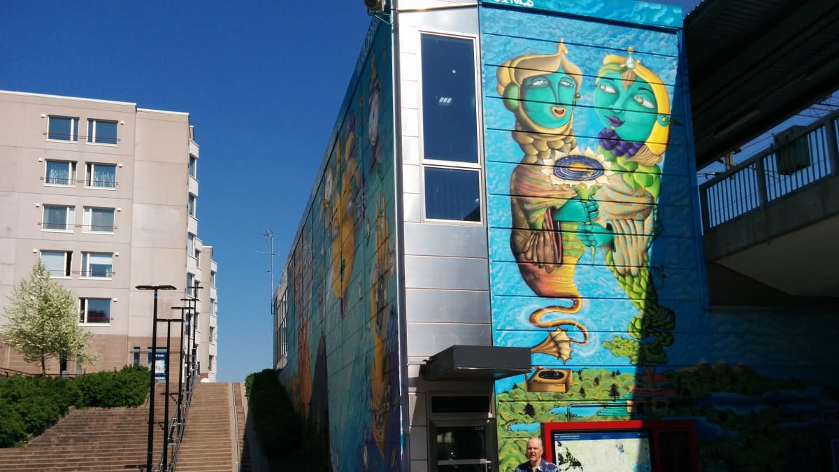 Myyrmäen asema. Seiniin maalattu katutaidetta. Rakennuksen edessä seisoo uuden paikallislehden Ari Martinaho.