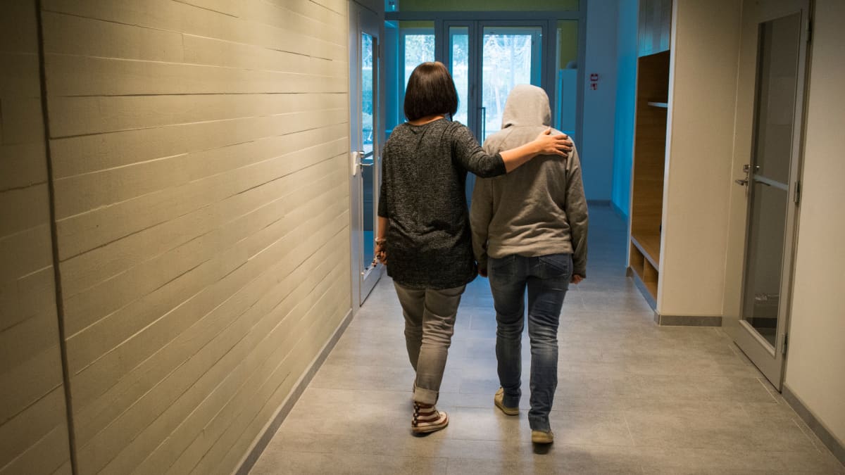 Huppariin pukeutunut nuori kävelee koulun käytävällä pää painuksissa, opettaja kulkee rinnalla käsi nuoren olkapäällä