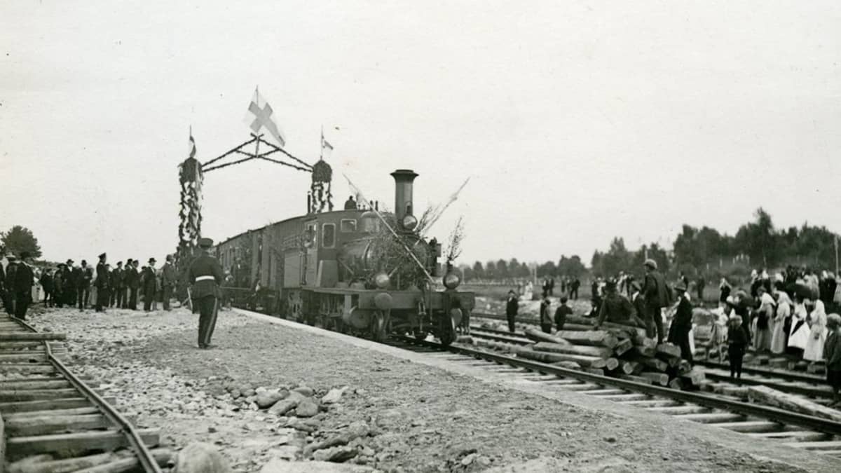 Hyöryjuna ajaa Suomen lipuin koristellun portin alitse. Ihmisiä on kerääntynyt katselemaan uutuutta.