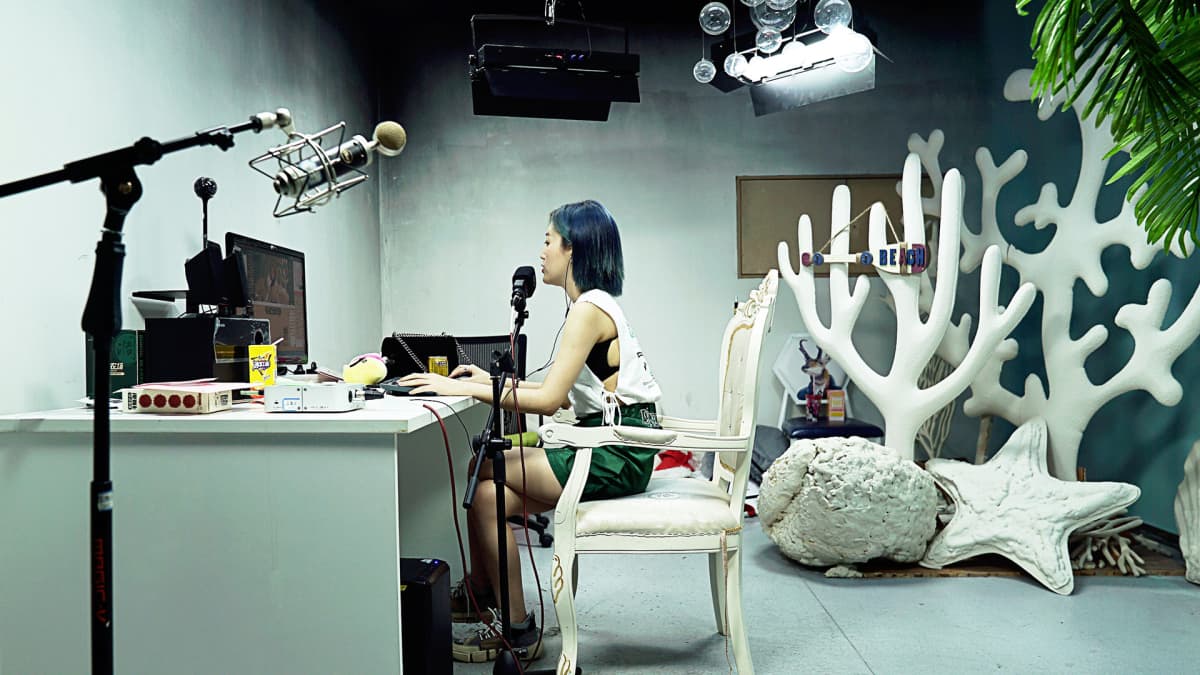 nainen istuu studiossa ympärillään kameroita, mikrofoneja ja lavasteita