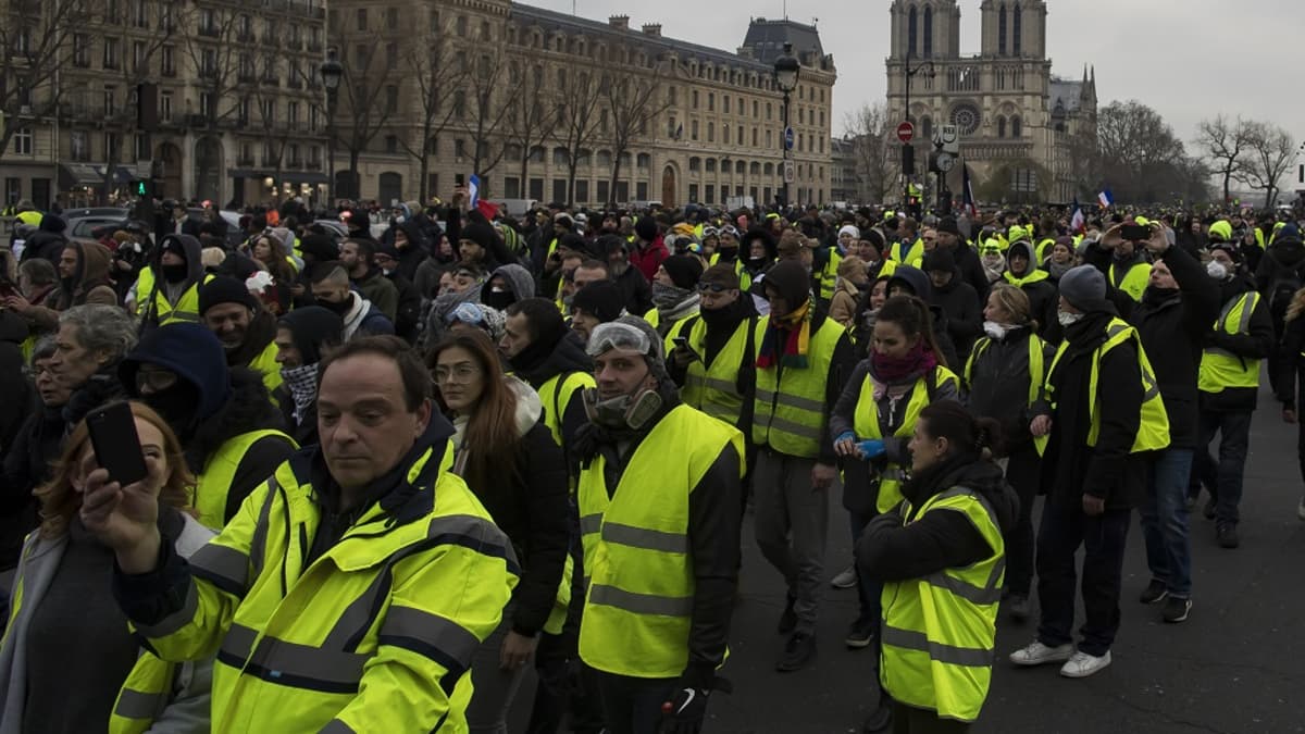 Keltaisiin huomioliiveihin pukeutuneet mielenosoittajat marssivat kadulla. Taustalla näkyy Notre Damen katedraali.