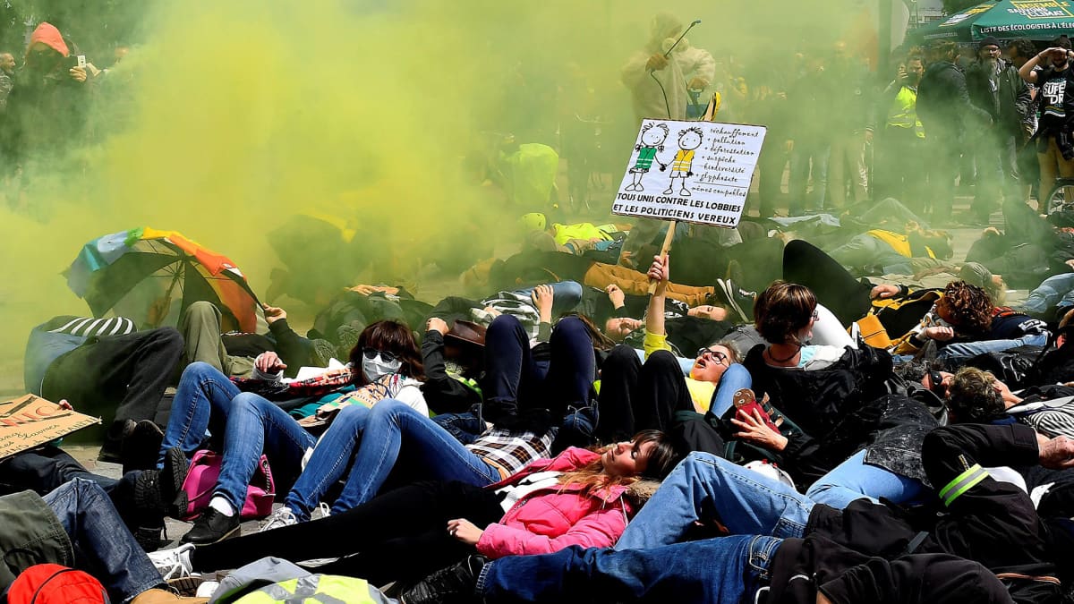 Keltaliivit ja ympäristöaktivistit protestoivat  Bayerin & Monsanto- yhtiötä vastaan 18. toukokuuta. Protestoijat makaavat maassa, keltaista väripuuteria leijuu ilmassa ja yksi protestoija esittää ruiskuttavasna kemikaalia maassa olevien päälle.