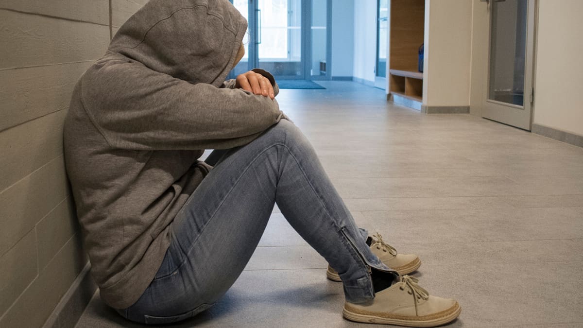 Huppariin kääriytynyt anonyymi nuori istuu koulun käytävällä.