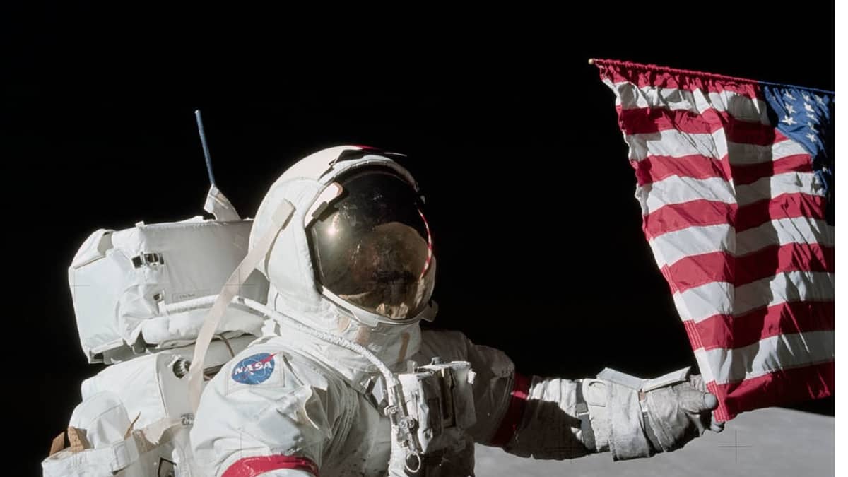 Astronautti pitelee kiinni lipun kulmasta. Taustalla avaruudessa häämöttää maapallo. 