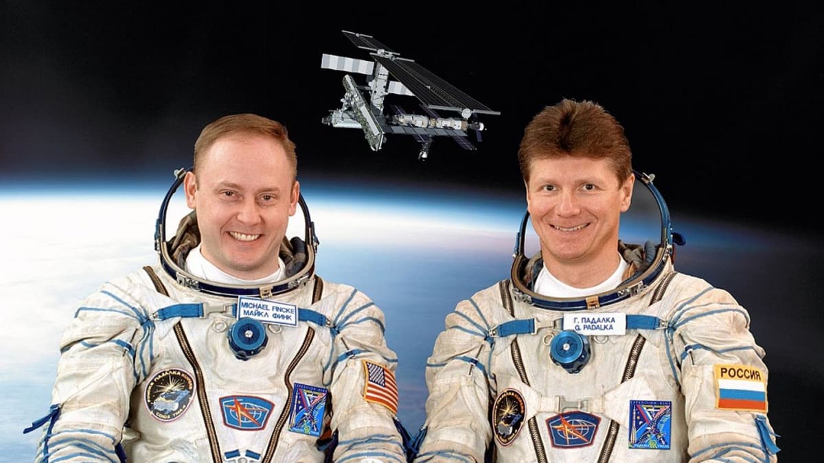 Kaksi avaruuspukuista miestä, taustalla kuva avaruusasemasta, alalaidassa 16 maan liput kertomassa, mistä maista ISS:llä on ollut väkeä.