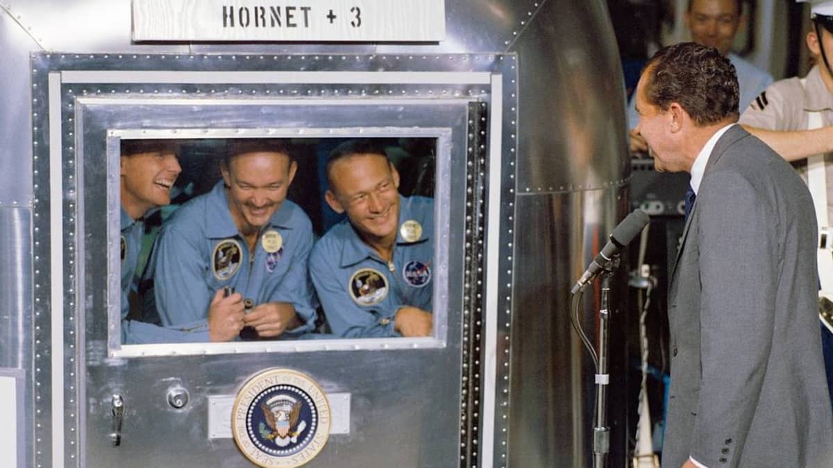 Arnstrong, Collins ja Aldrin kurkistevat hymyillen karanteenihuoneen ikkunasta, jonka edessä seisoo Nixon. 