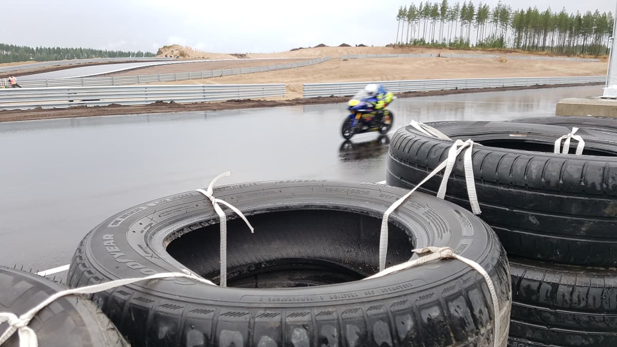 MotoGP-testikuljettaja sinikeltaisella moottoripyörällä Kymi Ringin märällä asfaltilla. Kuvattu radan reunan rengasvallin takaa.
