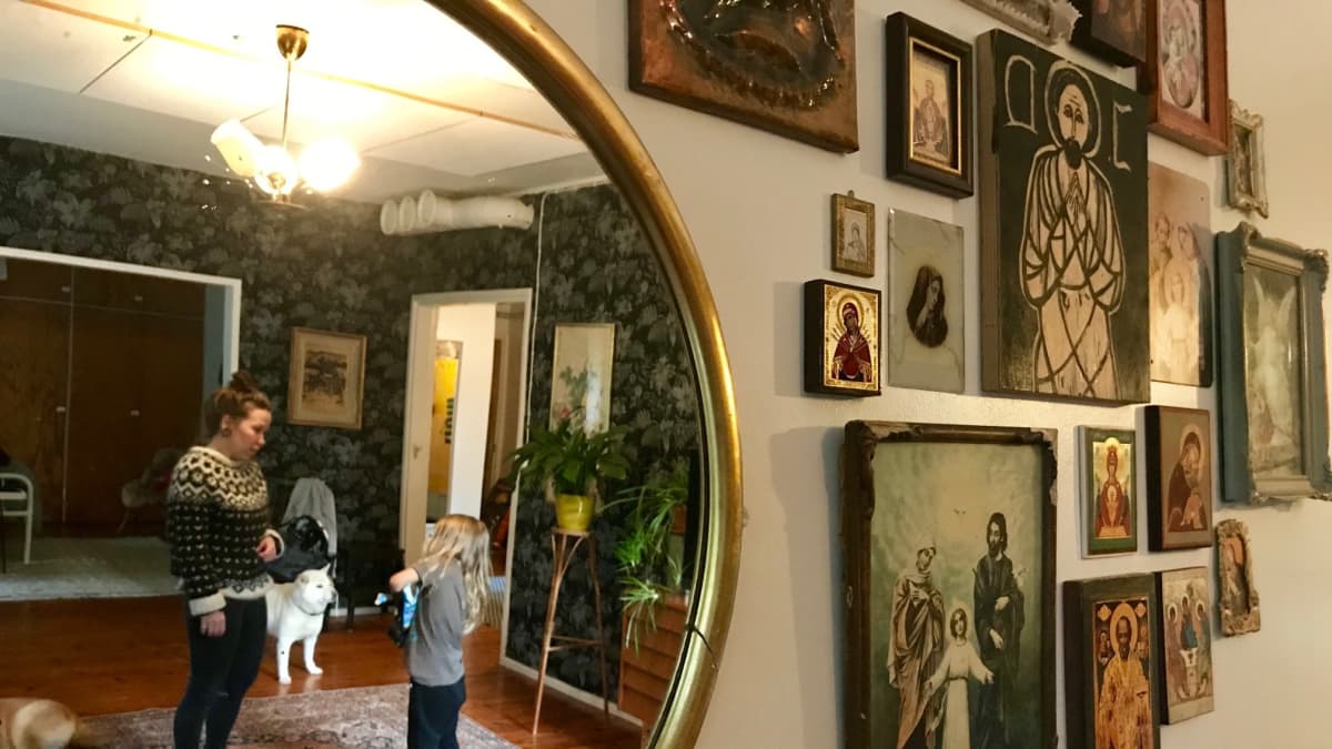 Ikoneja olohuoneen seinällä. Vieressä on pyöreä peili, josta kuvastuu nainen, lapsi ja kaksi koiraa. 