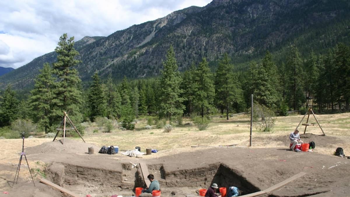 Arkeologeja kaivamassa kivirakennuksen raunioita. Taustalla korkeita vuoria. 