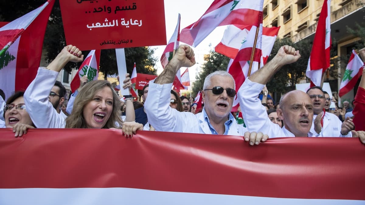 Etualalla kolme mielenosoittajaa punaisen lakanan takana. Heillä kaikilla on oikea käsi nyrkissä ja ylhäällä. Taustalla heilutetaan Libanonin lippuja. 