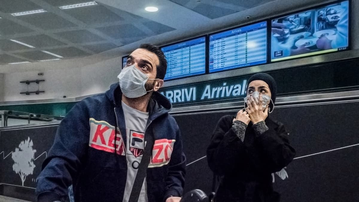 Mies ja nainen matkatavaroineen lentokentällä, molemmilla hengityssuojaimet. 