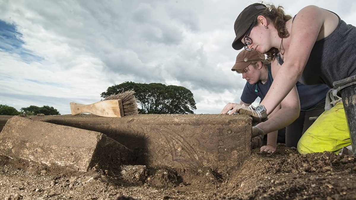 Kaksi nuorta arkeologia kaivaa maasta hautakiveä, jossa näkyy kaiverrusta. 