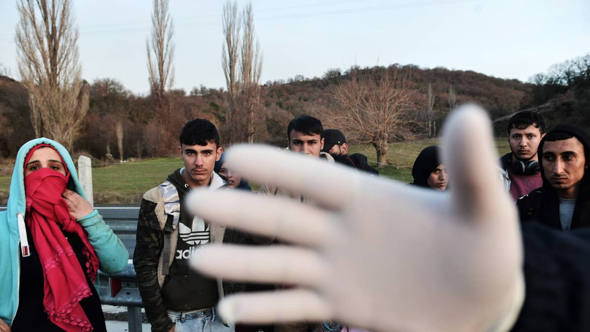 Kuvassa poliisin pidättämät siirtolaiset katsovat kohti kameraa, poliisin käsi on linssin edessä.