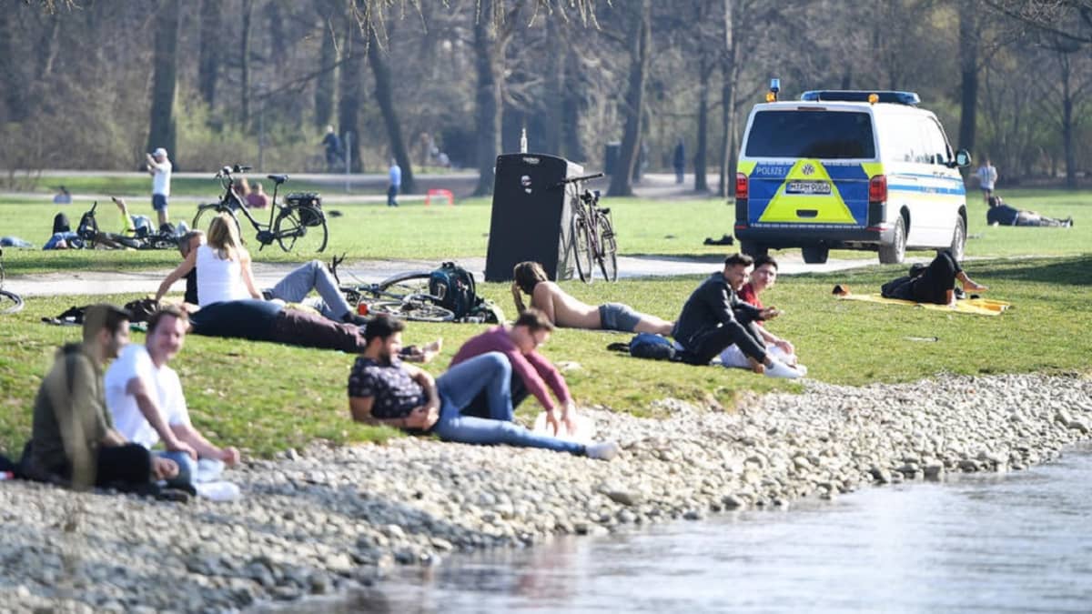 Kuvassa ihmisiä istumassa puistossa veden äärellä. Taustalla ohi ajava poliisiauto. 