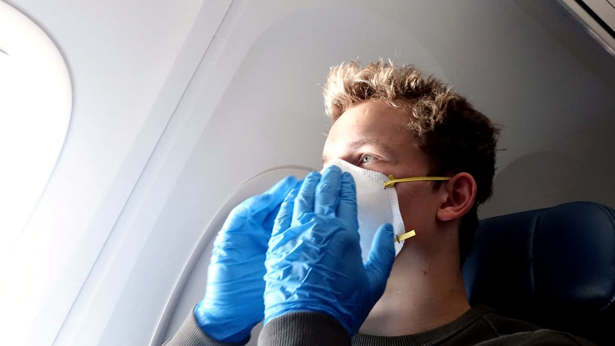 Hengityssuojaimeen ja suojakäsineisiin pukeutunut mies matkustaa lentokoneessa.