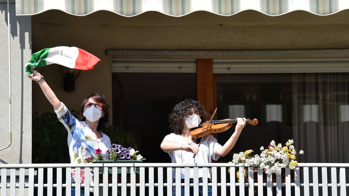 Nainen soittaa kasvomaskissa viulua parvekkeella, toinen nainen heiluttaa Italian lippua vieressä.