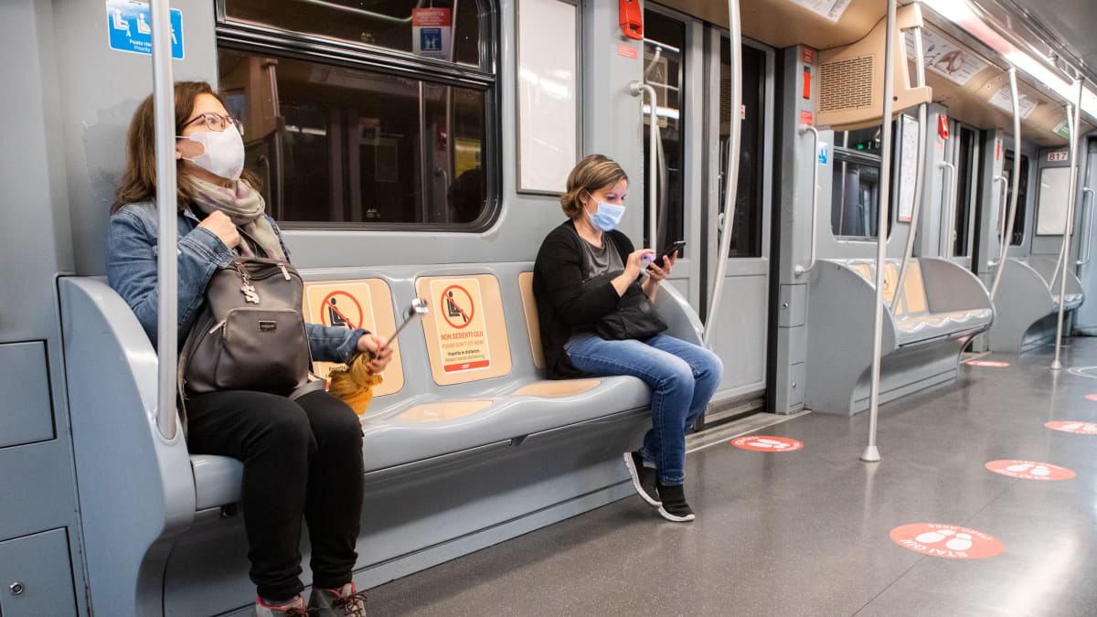 Matkustajat istuvat metrossa turvavälin päässä, turvaväliä osoittavia merkkejä Milanon metron lattiassa ja penkeillä