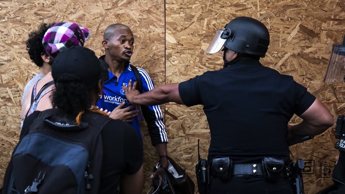 Kuvassa vasemmalla mustaihoinen mies. Selin kameraan oleva valkoihoinen poliisi on ojentanut kätensä kohti miestä. 