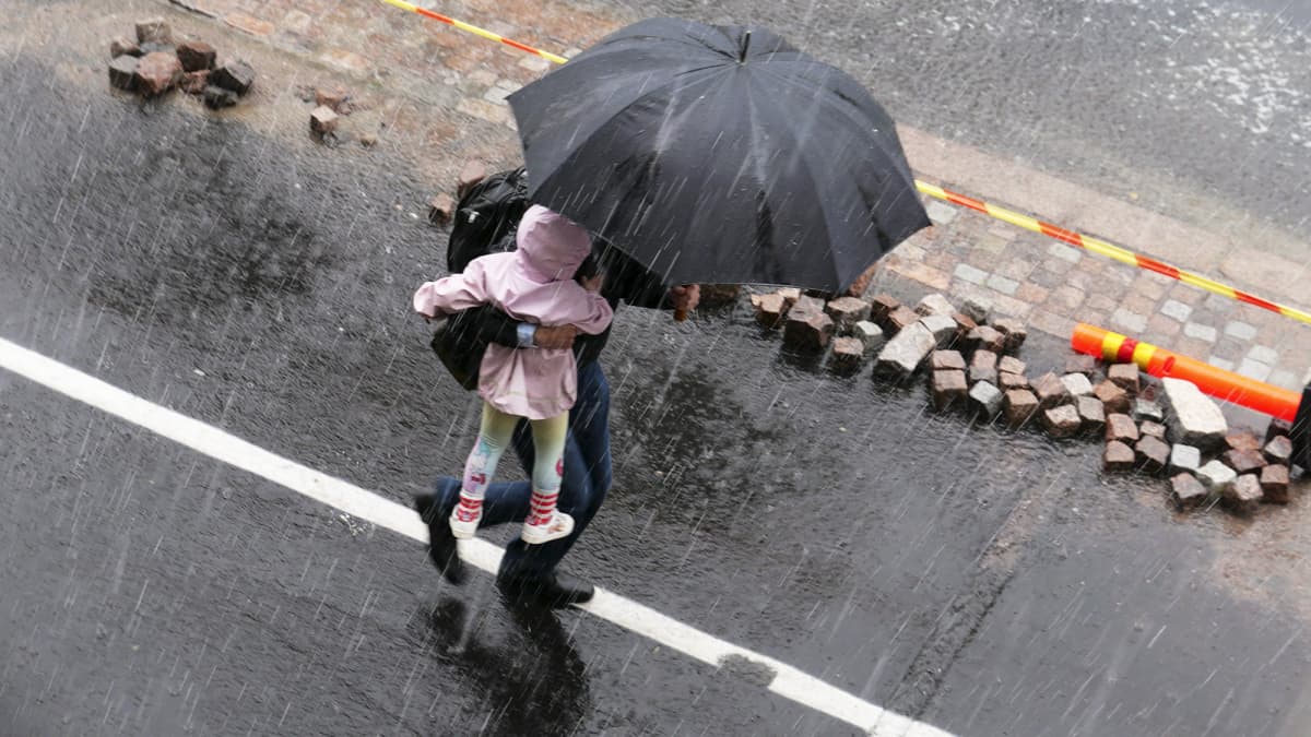 Mies kantaa lasta sateenvarjon alla rankkasateessa Helsingissä.