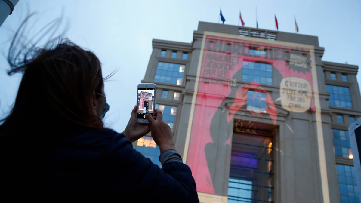 A woman takes pictures with her mobile phone as cartoons from French satirical newspaper Charlie Hebdo are projected onto buildings in central Montpellier in France, 21 October 2020. The cartoons of Prophet Mohammed were projected to pay tribute to the teacher Samuel Paty who was assassinated in Conflans-Sainte-Honorine. On 16 October French school teacher Samuel Paty was decapitated by 18-year-old attacker named Abdoulakh Anzorov who has been shot dead by policemen. Paty was a history teacher who had recently shown caricatures from Charlie Hebdo newspapers of the Prophet Mohammed in class. 