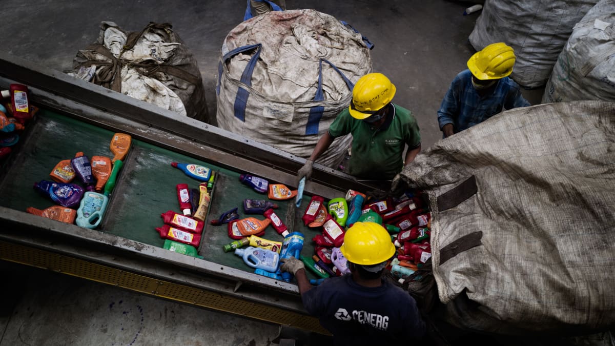 Kolme työntekijää kaataa suuresta säkistä käytettyjä muovipakkauksia liukuhihnalle.