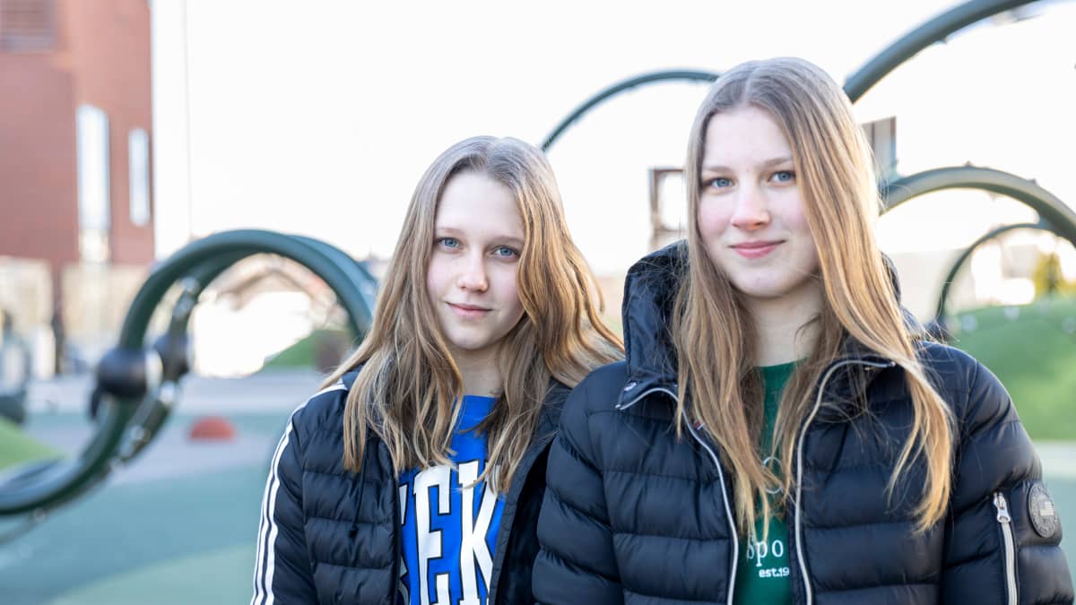 Kaksi teini-ikäistä tyttöä seisoo ulkona ja katsoo kameraan talvitakit päällään. Taustalla näkyy kiipeilytelinettä.