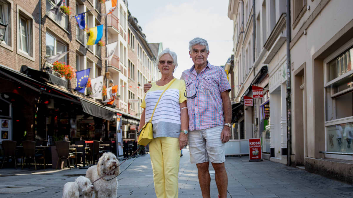 Harmaantunut pariskunta vanhhankaupungin aurinkoisella kadulla kahden valkoisen koiran kanssa. Mies pitää kättään vaimonsa olkapäällä.