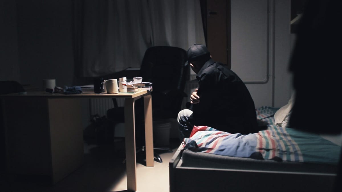 Mies istuu sängyllä pimeässä huoneessa, dokumenttielokuvan pressikuva.
