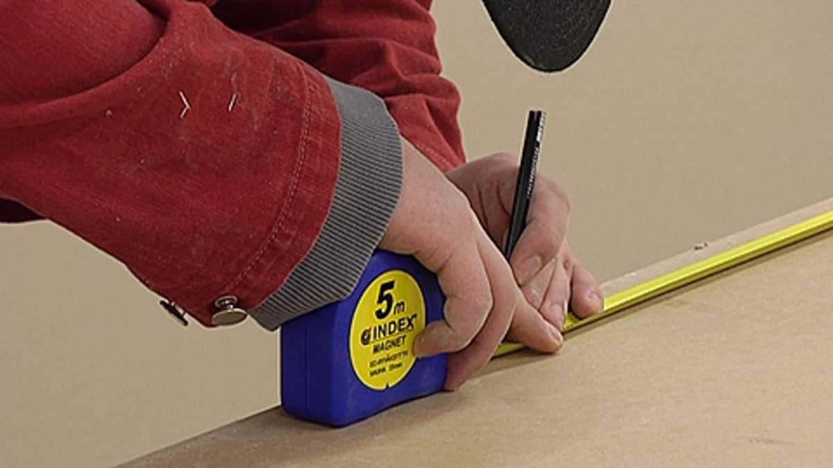 Rakennusmies mittaa levyä mittanauhalla ja tekee merkintää levyyn.