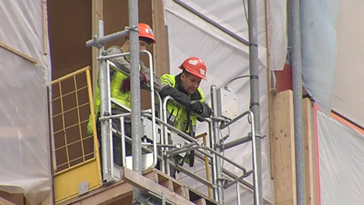 Kaksi rakennusmiestä nojaa kaiteeseen rakennuksen yläkerroksessa ja katsoo alas.