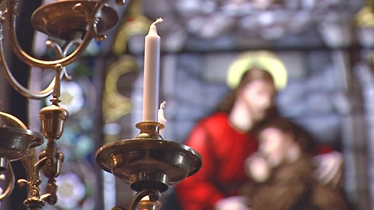 Polttamaton kynttilä on kiinnitetty koristeelliseen, moniosaiseen kynttilänjalkaan. Takana Jeesusta esittävä suurikokoinen lasimaalaus.