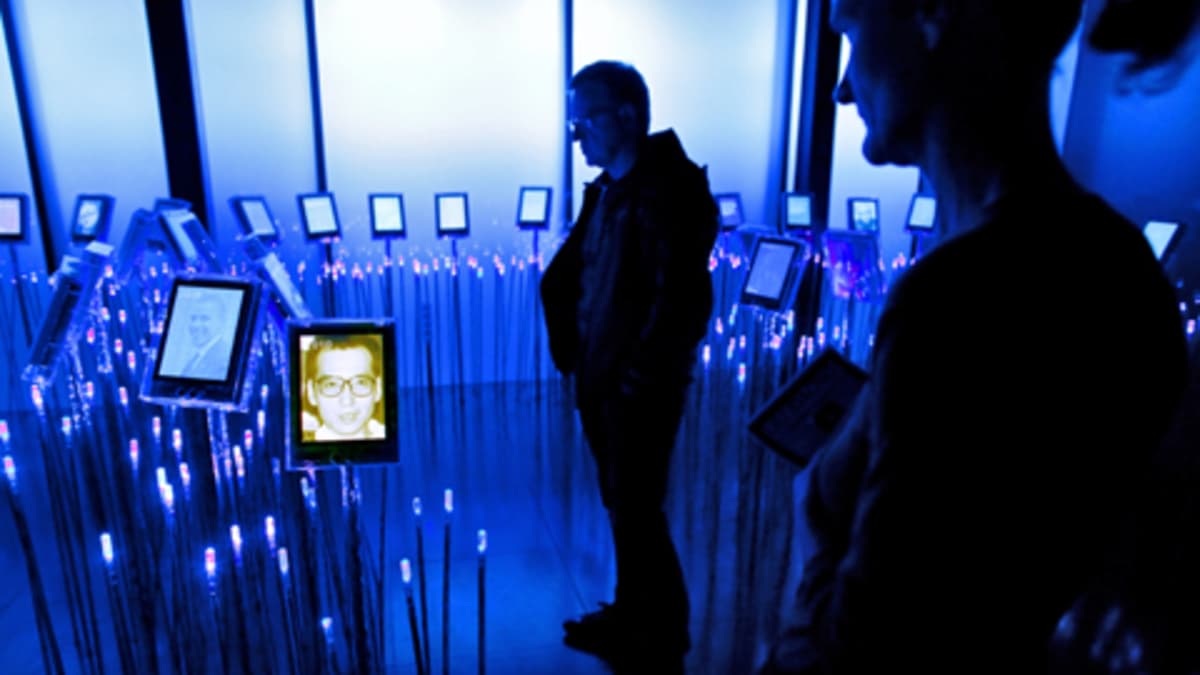 Sinisävyisessä huoneessa ihmiset katselevat Liu Xiaobon ja muiden aikaisempien rauhanpalkintojen saaneiden kuvia, jotka ovat jaluistoilla sähkökynttilöiden välissä.