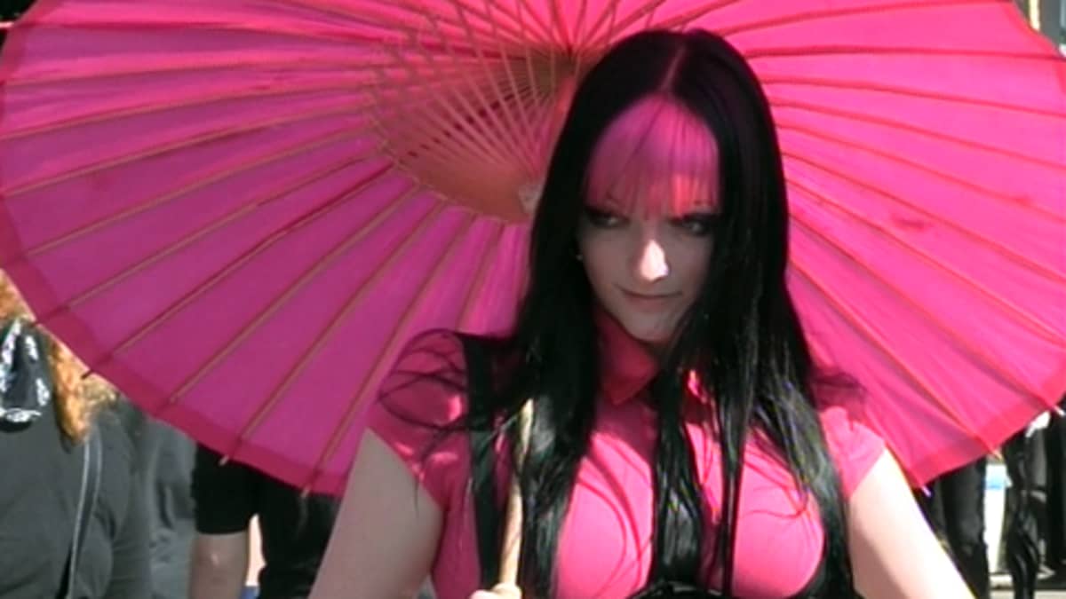 Nainen fuksian asussa vaaleanpunaisen päivävarjon alla. Fuksianpunaiset otsahiukset.