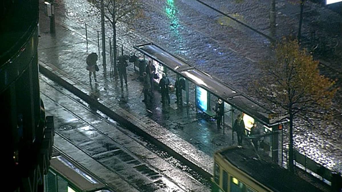 Ihmisiä sateessa raitiovaunupysäkillä.