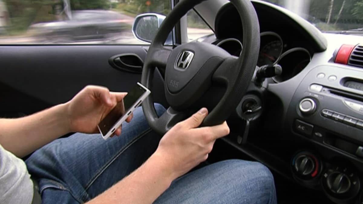 Mies ajaa autoa ja käyttää älypuhelinta.