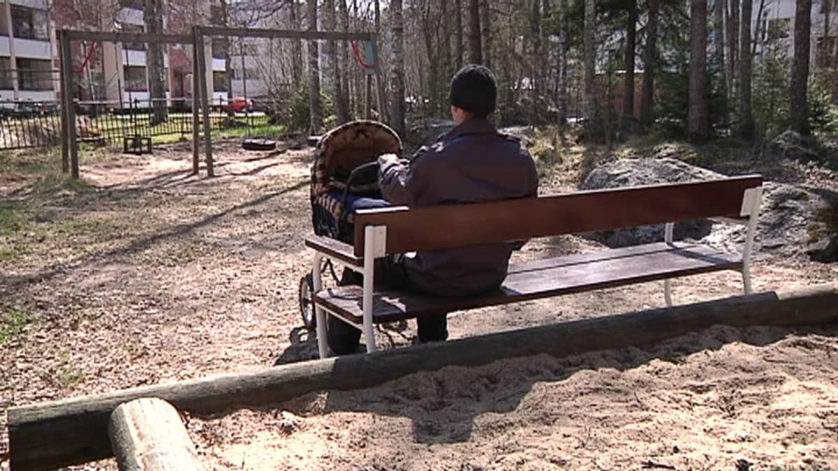 Kuvassa isä istuu puiston penkillä ja nukuttaa lasta vaunuissa.