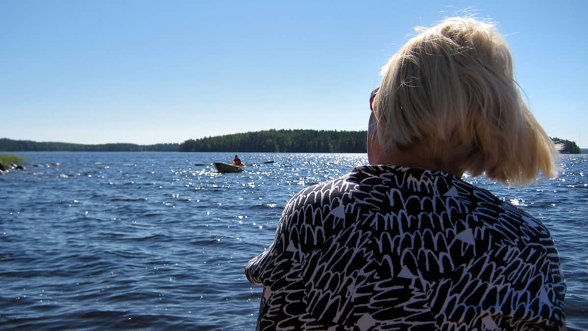 Nainen (selin) istuu järven rannalla, katselee järvellä olevaa soutuvenettä.