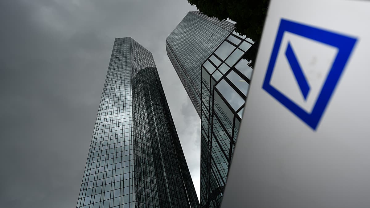 Kaksi pilvenpiirtäjää tummenevan taivaan alla Saksan Frankfurtissa. Etualalalla Deutsche Bankin logo.
