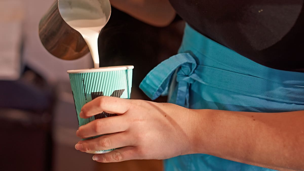 Kahvilatyöntekijä kaataa vaahdotettua maitoa pahviseen kahvinukiin.
