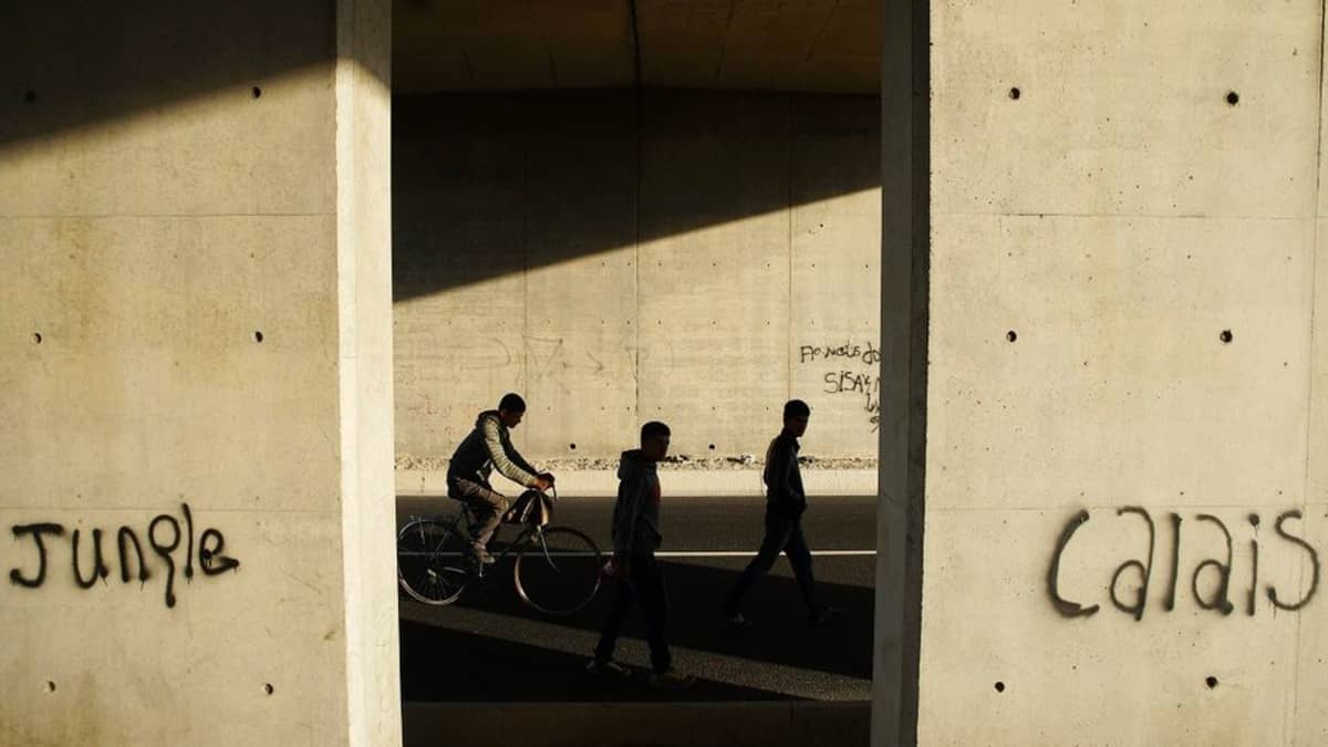 Käveleviä ja pyöräilevä miehiä betonipilarien väleissä. Pilarien graffiteissa lukee "Calais" ja "Jungle". 