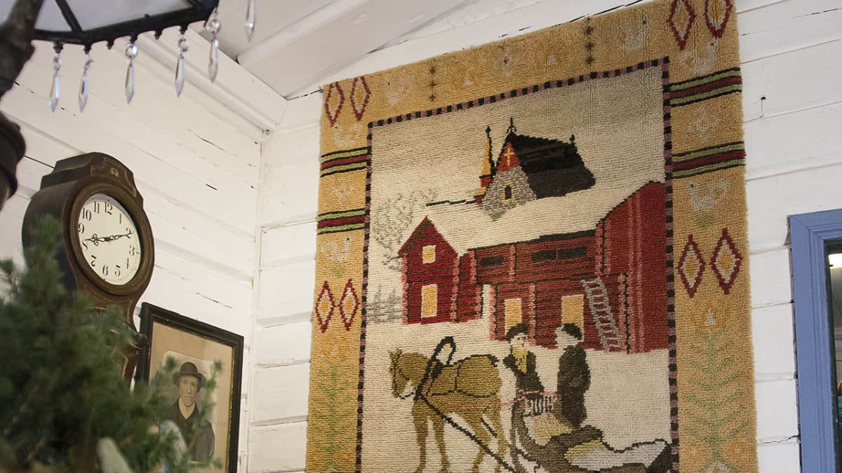 Seinällä olevan ryijy, jossa kuva kahdesta henkilöstä hevosreen kanssa lumisessa maisemassa. 